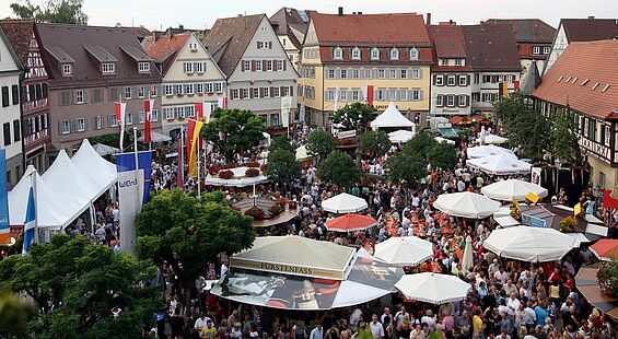 [Translate to Englisch:] Ein Luftbild vom Öhringer Weindorf ist zu sehen mit den Häusern rund um den Marktplatz, Menschen und großen Schirmen.